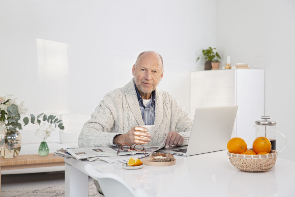 Mies istuu tietokoneen ääressä, kädessä on kahvikuppi ja pöydällä välipalaa.
