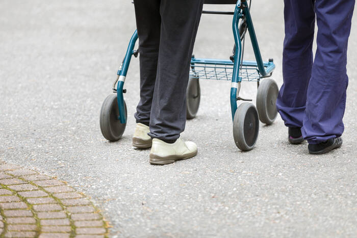 Kuvassa näkyvät ulkona kävelyllä olevan rollaattoria käyttävän vanhuksen jalat ja avustavan henkilön jalat.